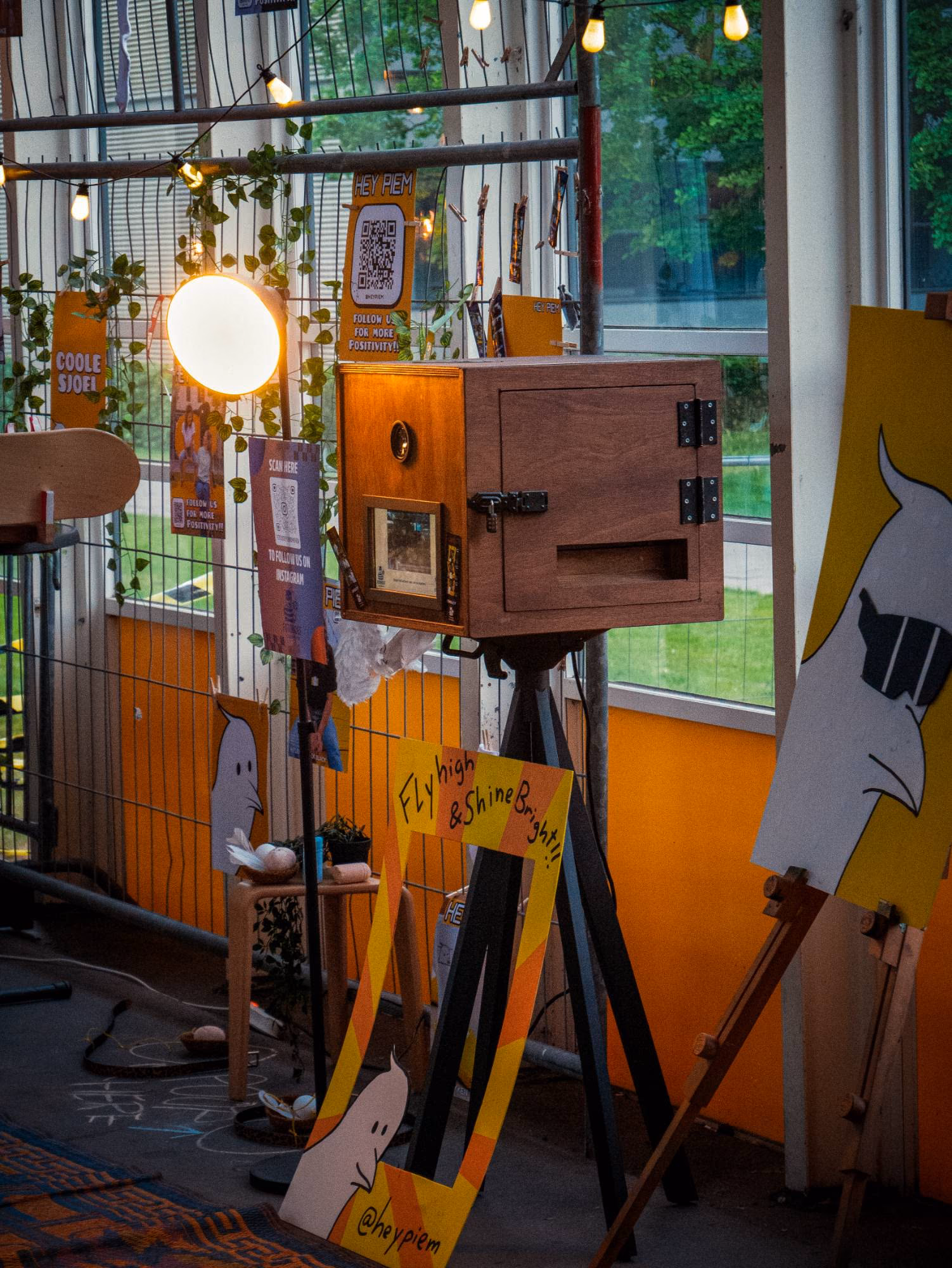 De houten photobooth op een zwart statief, met een lamp ernaast en een fotolijstbord ervoor.