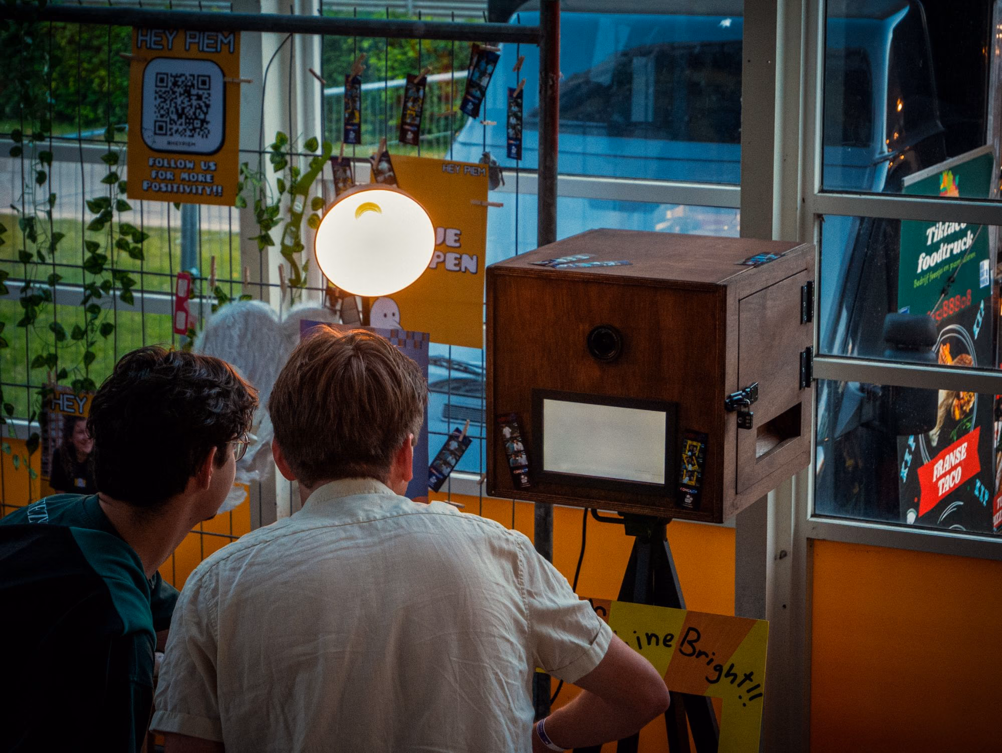 Twee gasten staan voor de houten photobooth naast een lamp.
