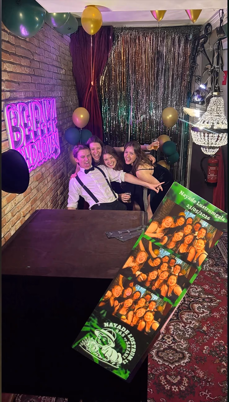 Gasten in galakleding poseren voor een glitterdoek, met hun groene fotostrip van het Nayade Lustrumgala.