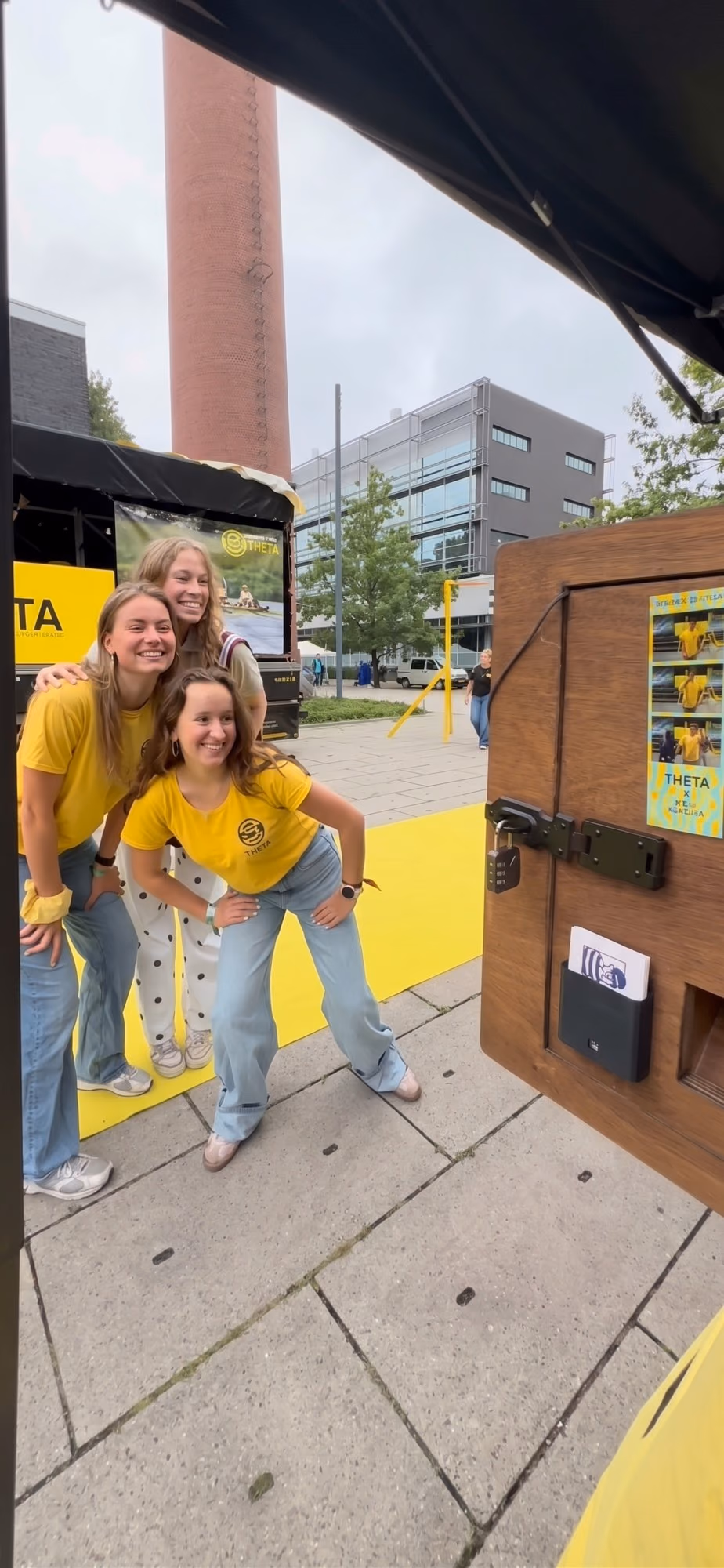 Drie studenten in gele shirts poseren lachend voor de photobooth op een campus.