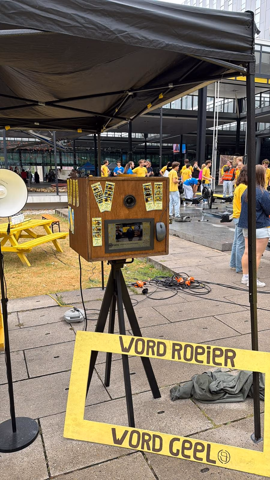 De photobooth op een statief onder een partytent, met fotostrips op de kast en een geel fotolijstbord ervoor.